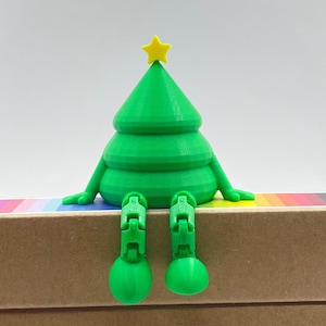 May include: A green 3D-printed Christmas tree with a yellow star, arms, and legs. The tree is sitting on a rainbow-striped surface, with flip-flop style legs. The figure is a festive decoration.