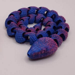 May include: A blue and pink articulated snake toy. The toy is made of connected, textured segments, with a gradient of blue and pink hues. The snake is coiled on a white surface.
