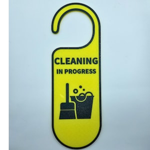 May include: A yellow door hanger with a black border and a curved top. The sign reads "CLEANING IN PROGRESS" above a black graphic of a broom and bucket. The sign is designed to hang on a door handle.