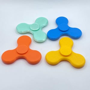 May include: Four brightly coloured fidget spinners in mint green, blue, orange, and yellow. Each has a three-lobed design with a central circular button. The spinners are arranged on a white surface.