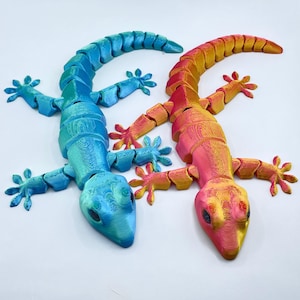 May include: Two articulated lizard toys. One is blue and turquoise, the other is pink, orange, and yellow. The lizards have segmented bodies, four legs with five toes each, and a long tail. The toys are on a white background.
