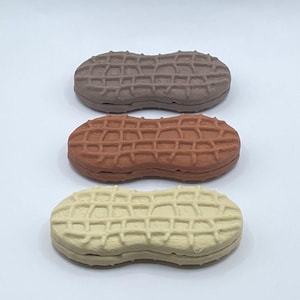 May include: Three peanut-shaped objects in shades of brown, orange, and cream. Each has a textured surface resembling a peanut shell. The objects are arranged vertically against a white background.