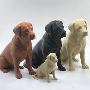 May include: Four Labrador Retriever dog figurines in shades of brown, black, and cream. The dogs are depicted in a seated pose, with a smaller figurine positioned in front. The figurines appear to be made from a solid material.