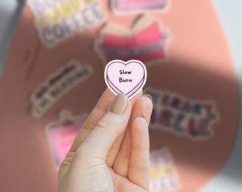 May include: A small, heart-shaped sticker with a light pink background and a white border, featuring the text "Slow Burn" in dark brown lettering. The sticker is held against a larger, oval-shaped pink board with other book-themed stickers.
