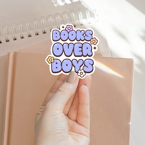 Può includere: Un adesivo blu chiaro con il testo "BOOKS OVER BOYS" in lettere bianche con contorno. L'adesivo ha piccoli disegni di fiori rosa, gialli e arancioni.