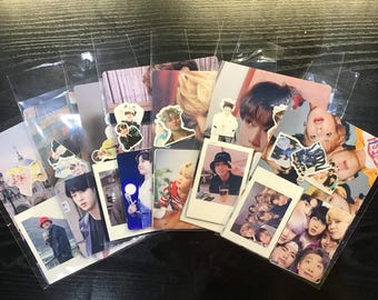 Op de afbeelding: Een verzameling fotokaarten met verschillende afbeeldingen van een populaire boyband. De kaarten zitten in doorzichtige plastic hoesjes en zijn op een donker oppervlak gerangschikt. Sommige kaarten hebben stickers erop.