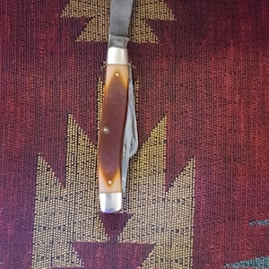 May include: A vintage pocket knife with a partially open blade. The handle is brown with a cream-colored border and silver accents. The knife is set against a patterned burgundy and gold background.