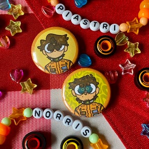 May include: Two circular yellow badges featuring cartoon illustrations of a person in a racing suit. The badges are surrounded by beaded bracelets with white letter beads spelling "PIASTRI" and "NORRIS", plus star and heart-shaped beads.