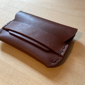 May include: A brown leather wallet with a flap closure. The wallet has a minimalist design and is stitched with dark brown thread.