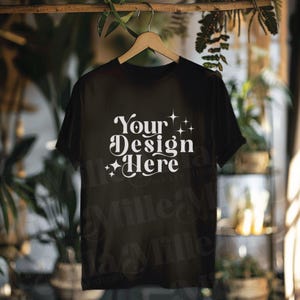 May include: Black t-shirt mockup with the text "Your Design Here" in white on the front. The shirt is hanging on a wooden hanger against a blurred background.