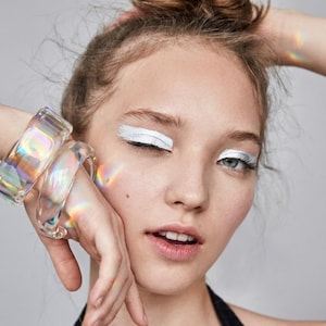 Peut inclure: Deux bracelets joncs transparents, épais et arrondis, dotés d'une finition holographique irisée qui reflète les couleurs de l'arc-en-ciel. Les bracelets sont portés au poignet et captent la lumière pour afficher des spectres changeants.