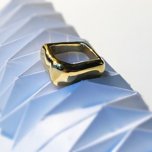 May include: A gold-colored, square-shaped ring with a smooth, slightly rounded surface. The ring is sitting on a white, folded paper background.