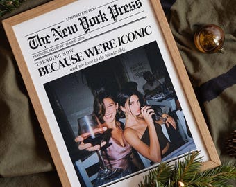 May include: A framed print resembling a newspaper front page, featuring the headline "BECAUSE WE'RE ICONIC." The image shows two women holding drinks. The frame is light brown, set against a green cloth with black stripes, pine needles, and ornaments.