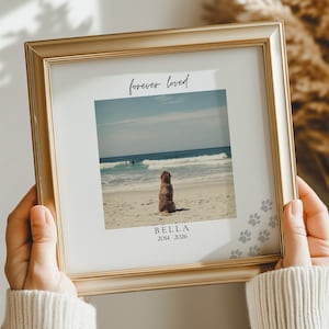 May include: A square gold-toned picture frame containing a photograph of a dog sitting on a sandy beach facing the ocean. The print features the text "forever loved" at the top and "BELLA 2014 - 2026" at the bottom, with grey paw prints.