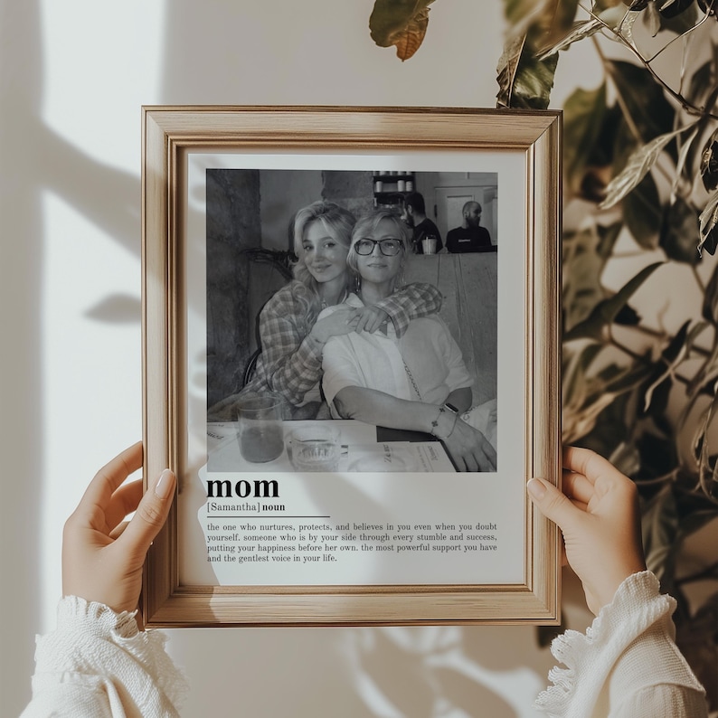 May include: A framed black and white photograph featuring two women embracing. The frame is a light brown color. Below the photograph is the word "mom" with a definition. The photograph is held by a person.