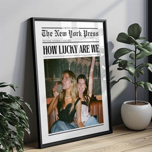 May include: A framed print designed to look like a newspaper front page. The headline reads "HOW LUCKY ARE WE" above a photo of two women. The newspaper title is "The New York Press" with the date "SATURDAY 6 JULY 2000X".