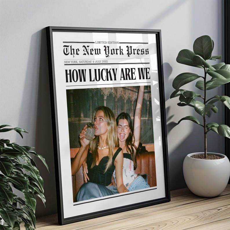 May include: A framed print designed to look like a newspaper front page. The headline reads "HOW LUCKY ARE WE" above a photo of two women. The newspaper title is "The New York Press" with the date "SATURDAY 6 JULY 2000X".