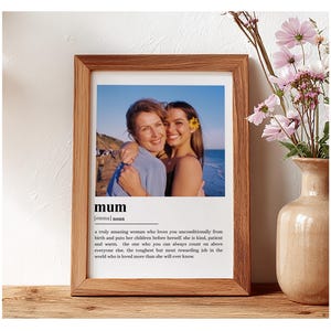 May include: A framed print with a photograph of two women embracing, with the word "mum" and a definition below. The frame is brown, and the print is on a wooden surface. A vase of pink flowers is to the right.