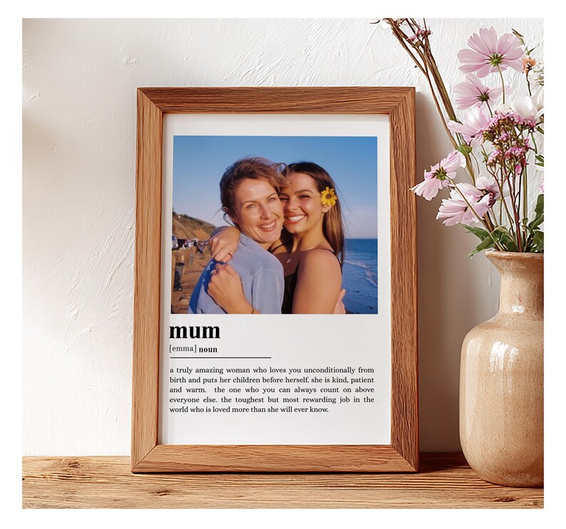 May include: A framed print with a photograph of two women embracing, with the word "mum" and a definition below. The frame is brown, and the print is on a wooden surface. A vase of pink flowers is to the right.