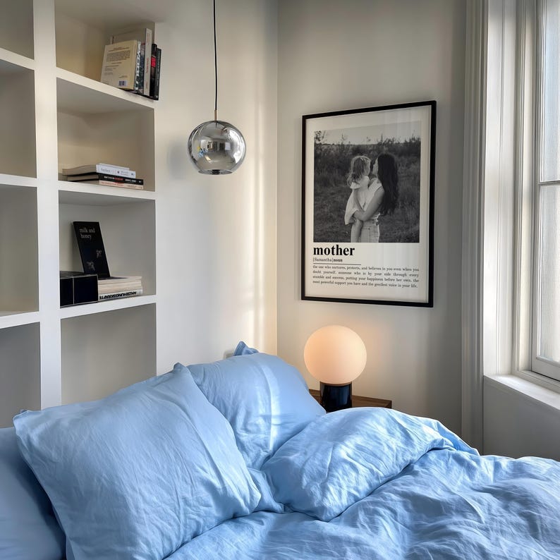 May include: A bedroom scene featuring a bed with light blue bedding, a framed black and white print with the word "mother", and a spherical pendant light. A small, round lamp sits on a nightstand. Bookshelves are visible in the background.
