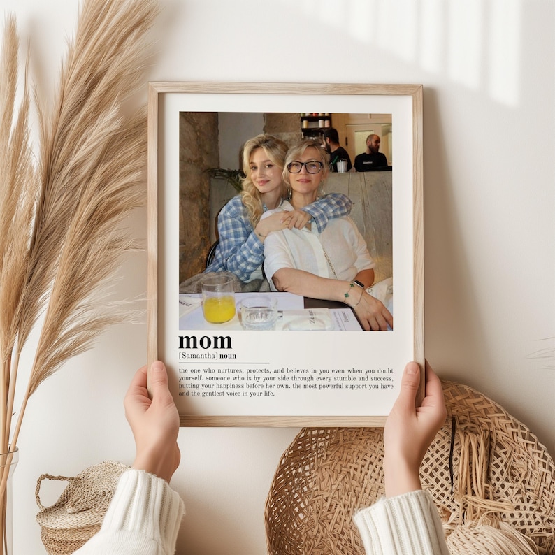 May include: A framed print featuring a photograph of two women embracing. The print includes the word "mom" with a definition below. The frame is light-colored, and the background includes dried plants and a woven basket.