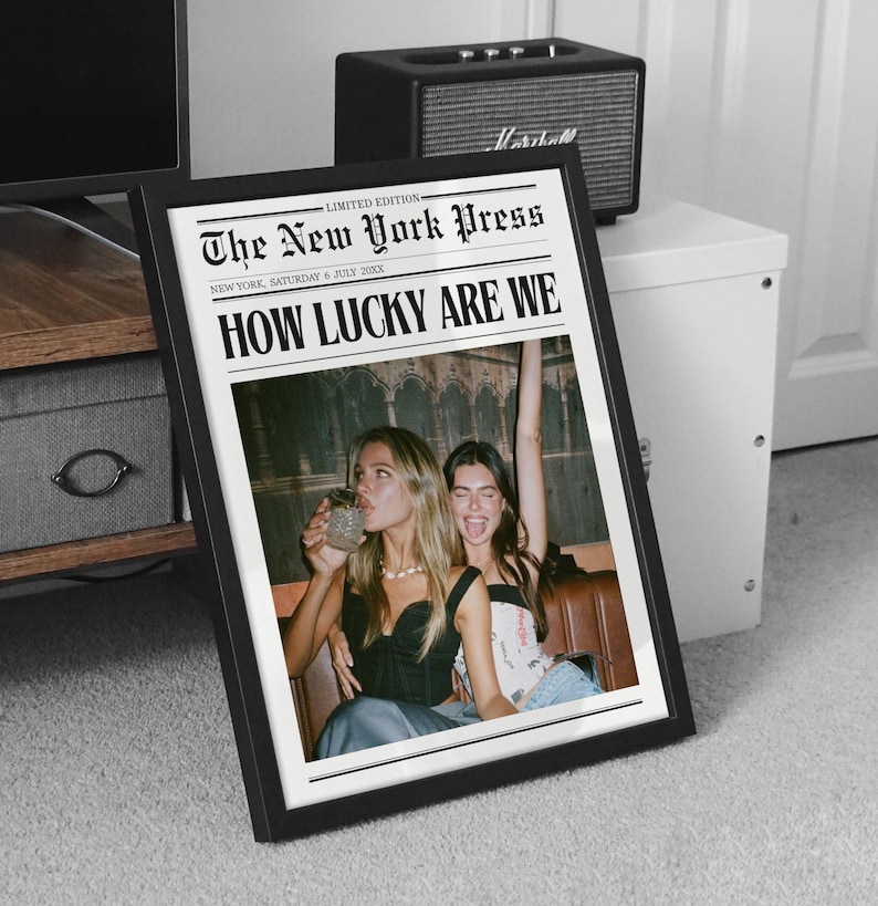 May include: A framed print featuring a newspaper-style design with the headline "HOW LUCKY ARE WE." The text "The New York Press" and "LIMITED EDITION" are also visible. The image inside the frame shows two women. A black speaker is in the background.