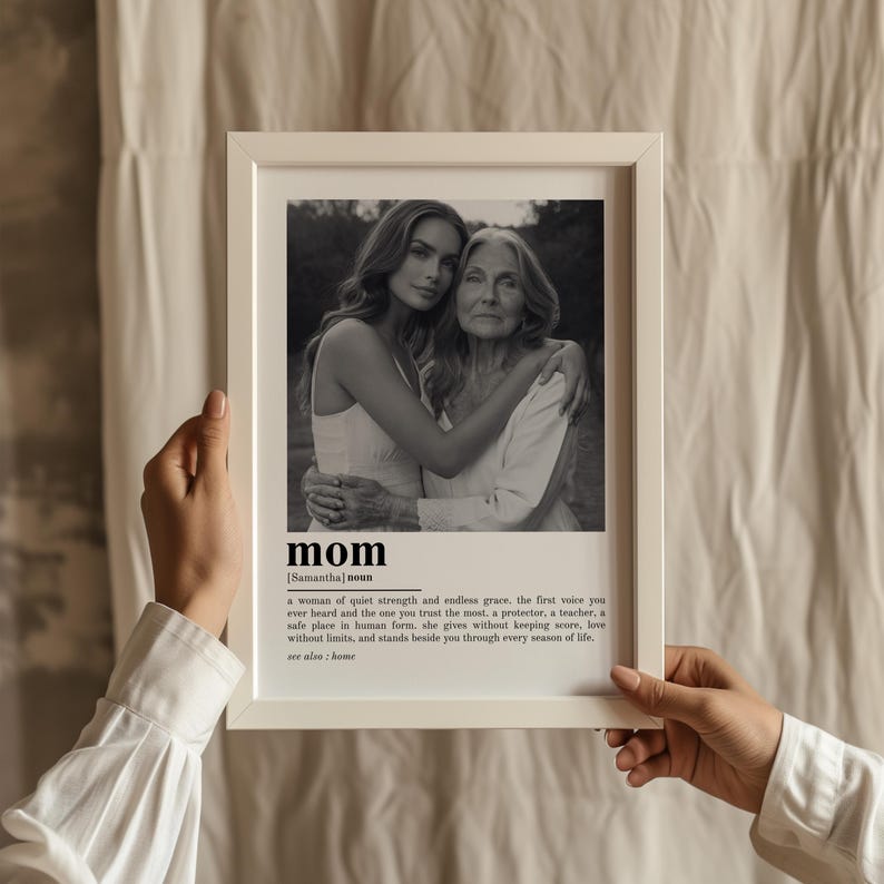 May include: A framed black and white photograph featuring two women embracing. The word "mom" is printed in bold letters at the bottom, along with a definition. The frame is white and held by a person.