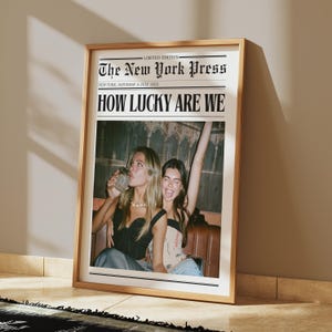 May include: Framed print of a newspaper front page titled "The New York Press" with the headline "HOW LUCKY ARE WE." The image features two women, one drinking from a glass and the other with her arm raised. The frame is a light wood color.