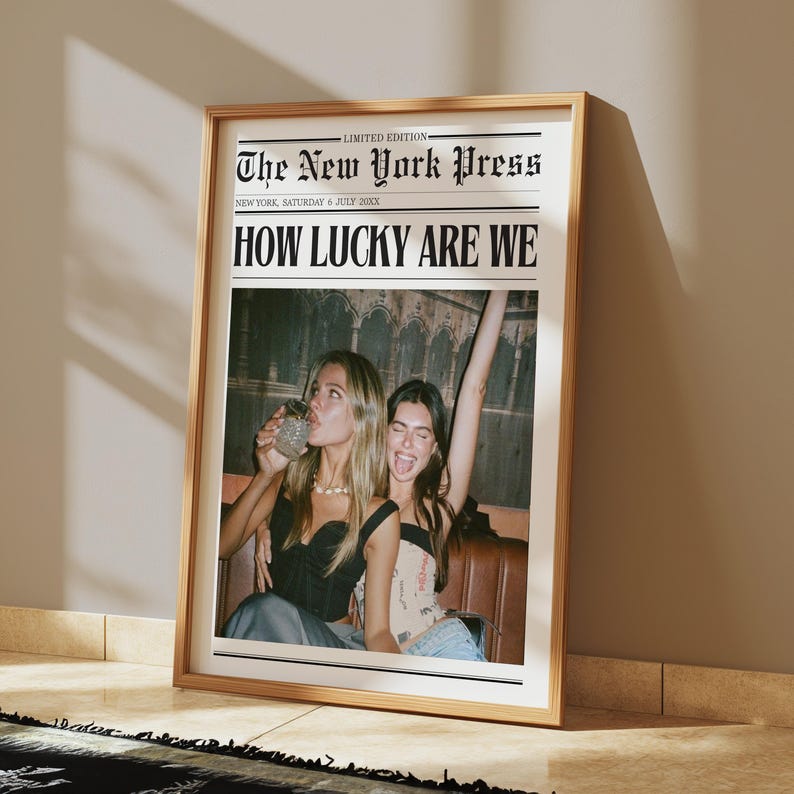 May include: Framed print of a newspaper front page titled "The New York Press" with the headline "HOW LUCKY ARE WE." The image features two women, one drinking from a glass and the other with her arm raised. The frame is a light wood color.
