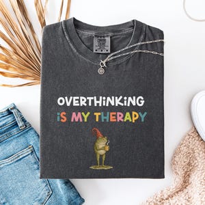 May include: Dark gray t-shirt with the text "OVERTHINKING IS MY THERAPY" in white and colorful letters. A cartoon frog wearing a red hat and holding a mug is printed below the text. The shirt is paired with blue jeans and white sneakers.