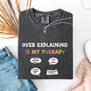 May include: Dark gray t-shirt with the text "OVER EXPLAINING IS MY THERAPY" in white and colorful letters. The shirt has speech bubbles with phrases like "I have a huge bumd on my..." and "You get it? Because..." laid out on a white surface.