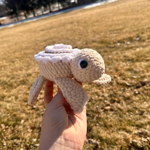 Puede incluir: Una tortuga de ganchillo beige con un caparazón blanco. La tortuga tiene ojos negros y se sostiene en una mano.