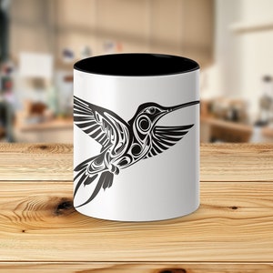 Puede incluir: Una taza de cer&aacute;mica blanca con un borde y un asa negros. La taza presenta una ilustraci&oacute;n estilizada en blanco y negro de un colibr&iacute; en vuelo.