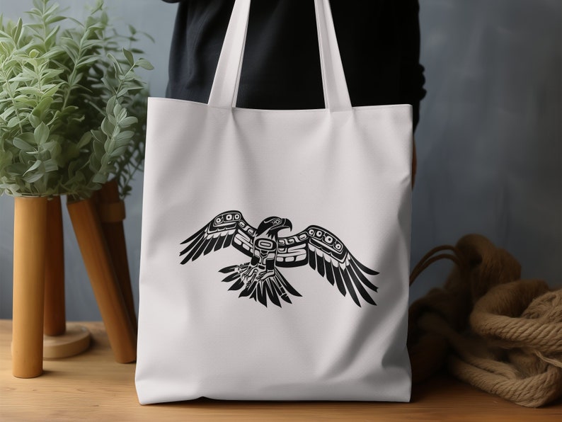 K&ouml;nnte beinhalten: Wei&szlig;e Canvas-Tragetasche mit einer stilisierten Schwarz-Wei&szlig;-Illustration eines Vogels mit ausgebreiteten Fl&uuml;geln. Der Vogel ist im traditionellen indianischen Stil gehalten.