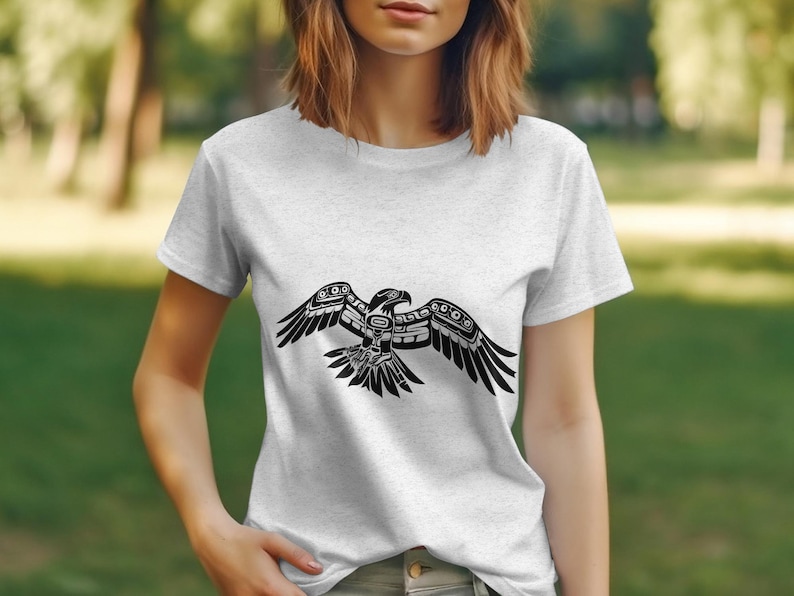 K&ouml;nnte beinhalten: Ein wei&szlig;es T-Shirt mit einem schwarzen, stilisierten Design eines Vogels mit ausgebreiteten Fl&uuml;geln. Der Vogel ist im traditionellen indianischen Stil gehalten.
