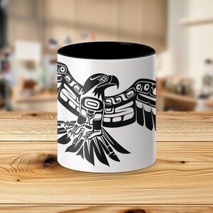 K&ouml;nnte beinhalten: Eine wei&szlig;e Keramiktasse mit schwarzem Rand und Henkel. Die Tasse zeigt eine stilisierte Schwarzwei&szlig;illustration eines Adlers mit ausgebreiteten Fl&uuml;geln. Der Adler ist im traditionellen Stil der amerikanischen Ureinwohner gehalten.