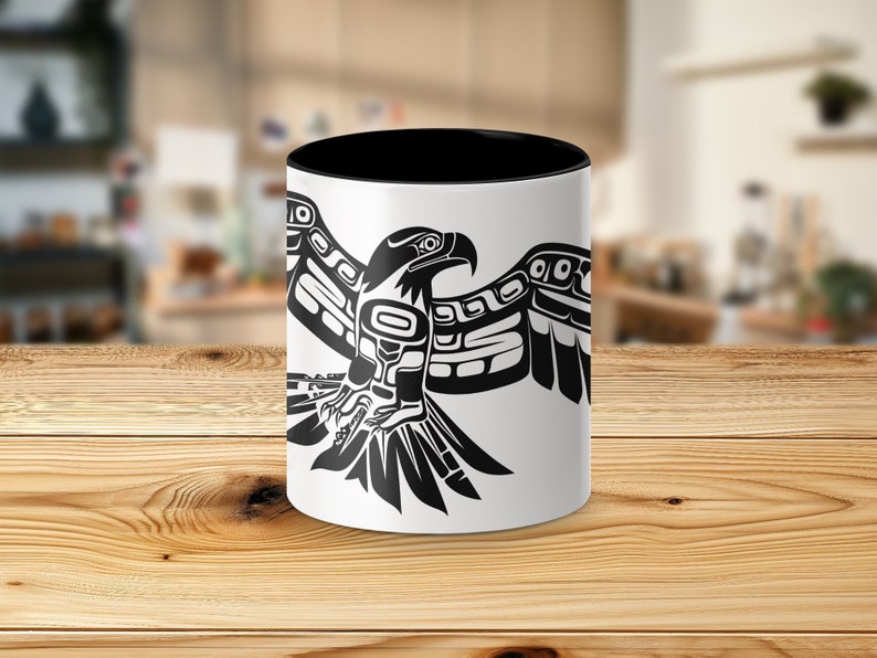 K&ouml;nnte beinhalten: Eine wei&szlig;e Keramiktasse mit schwarzem Rand und Henkel. Die Tasse zeigt eine stilisierte Schwarzwei&szlig;illustration eines Adlers mit ausgebreiteten Fl&uuml;geln. Der Adler ist im traditionellen Stil der amerikanischen Ureinwohner gehalten.