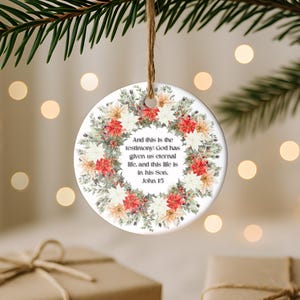Könnte beinhalten: Runder Weihnachtsornament aus Keramik mit einem floralen Kranz in Rot-, Weiß- und Pfirsichtönen. Das Ornament trägt ein Zitat aus Johannes 1:5. Eine Schlaufe aus Jute-Schnur ist zum Aufhängen befestigt.