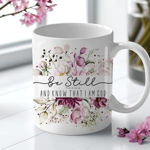 May include: White ceramic mug with floral design in shades of pink, purple, and green. The mug features the text "Be Still and Know That I Am God" in black script. A decorative item for the home.