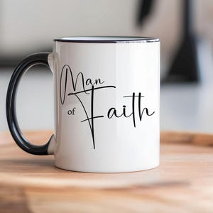May include: White ceramic mug with a black handle and rim. The mug features the words "Man of Faith" in elegant black script. The mug is sitting on a wooden tray.