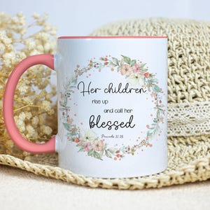 May include: White ceramic mug with a pink rim and handle. The mug features a floral wreath with the text "Her children rise up and call her blessed Proverbs 31:28".