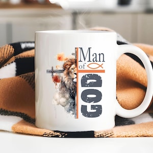 May include: A white ceramic mug featuring a watercolor-style illustration of a lion's face on the left side, partially obscured by a vertical cross. The text "Man of GOD" is printed in dark gray and orange, including a fish symbol.