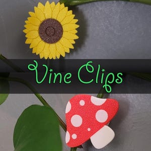 May include: Two plant clips, one shaped like a sunflower with yellow petals and a brown centre, and the other shaped like a red and white mushroom with white dots. The text "Vine Clips" is written in green letters on a black background.