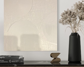 May include: A white abstract painting with a textured surface featuring concentric circles and lines. The painting is hanging on a white wall above a dark wood table with a black vase and dried flowers.