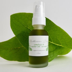 May include: A glass bottle of comfrey & castor oil serum, labeled "Midheaven Body", with a green liquid inside. The bottle is sitting on a green leaf.