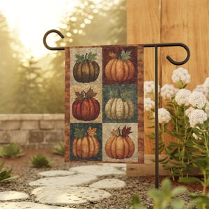 May include: A decorative garden flag featuring a patchwork design of six pumpkins with green leaves. The pumpkins are in shades of brown, orange, and cream, set against a background of autumnal colors. The flag is displayed on a black metal garden stake.