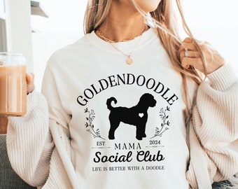 Può includere: Felpa bianca con testo nero che recita "Goldendoodle Mama Social Club Est. 2024 Life is better with a doodle." Il testo è circondato da una ghirlanda di foglie. Una silhouette nera di un cane Goldendoodle è al centro della ghirlanda.
