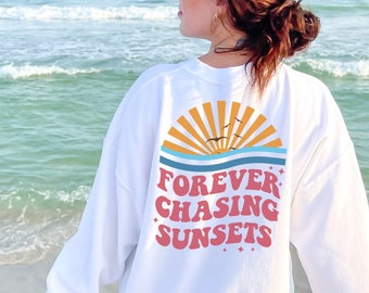 Puede incluir: Sudadera blanca con un gráfico de una puesta de sol y el texto "FOREVER CHASING SUNSETS" en rosa y amarillo.