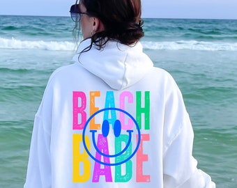 Puede incluir: Sudadera blanca con un gráfico de colores arcoíris que dice "Beach Babe" con una carita sonriente azul en el medio.