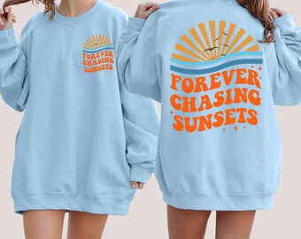 Puede incluir: Una sudadera azul claro con el texto "FOREVER CHASING SUNSETS" en naranja en la parte delantera y trasera. El diseño de la parte trasera presenta una puesta de sol estilizada con pájaros volando sobre el océano.